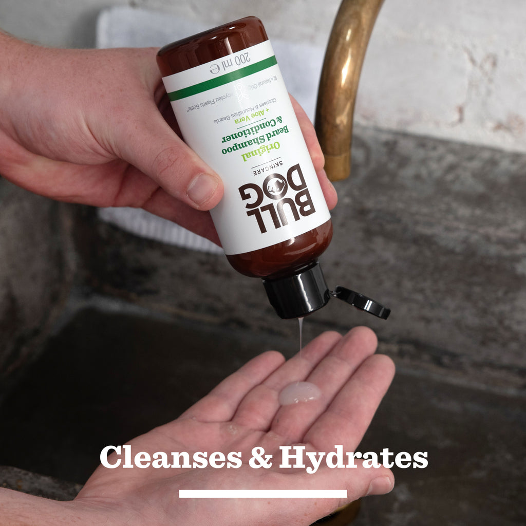 A person squeezes Bull Dog's Original 2-in-1 Shampoo & Conditioner from their Original Beard Care Kit onto their palm over a sink. Text at the bottom reads: Cleanses & Hydrates for effortless beard grooming.