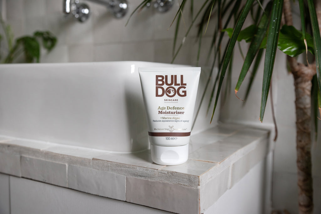 A tube of Age Defence Moisturiser for men rests on a white tiled sink, with a green plant and faucet in the background, emphasizing its natural origin ingredients.