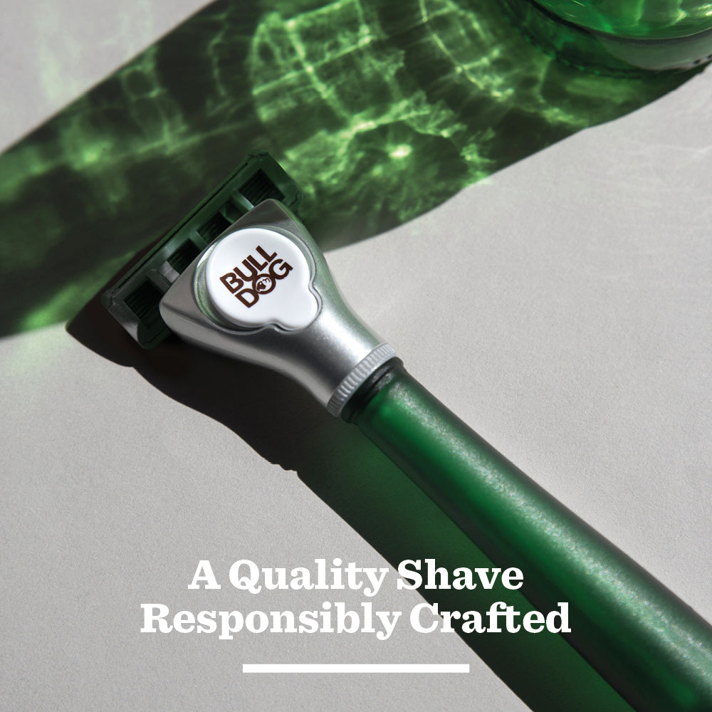 The Original Glass Razor with a green handle rests diagonally on a light surface with green reflections, showcasing its plastic-free packaging and the text: A Quality Shave Responsibly Crafted.