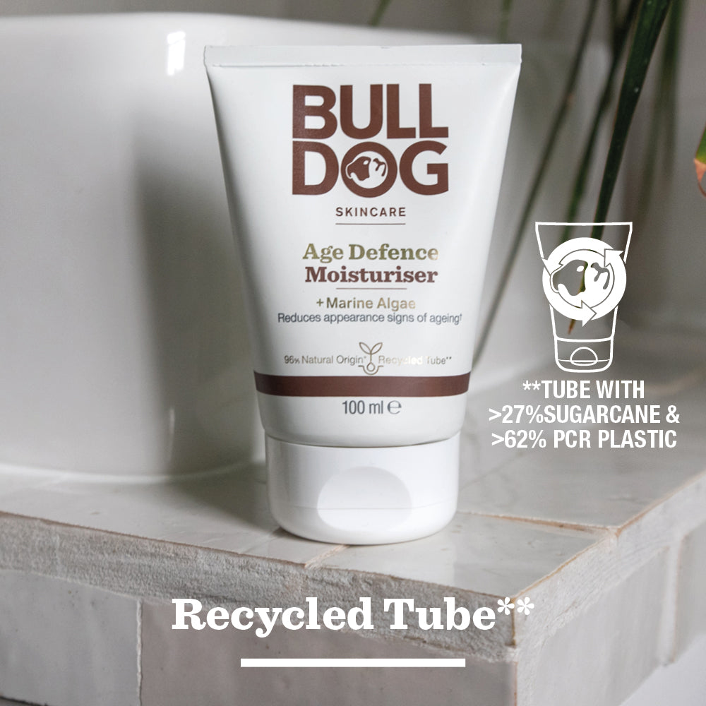 A tube of Age Defence Moisturiser for men rests on a white bathroom surface. Text states it’s made with over 27% sugarcane, 62% PCR plastic, uses natural origin ingredients, and is labeled as a Recycled Tube.