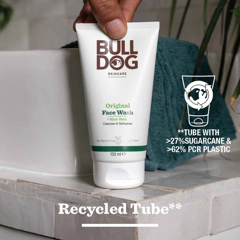 A hand holds a tube of Original Face Wash above a bathroom sink. The text notes its natural origin ingredients, made with over 27% sugarcane and 62% PCR plastic. Recycled Tube appears at the bottom of the image.