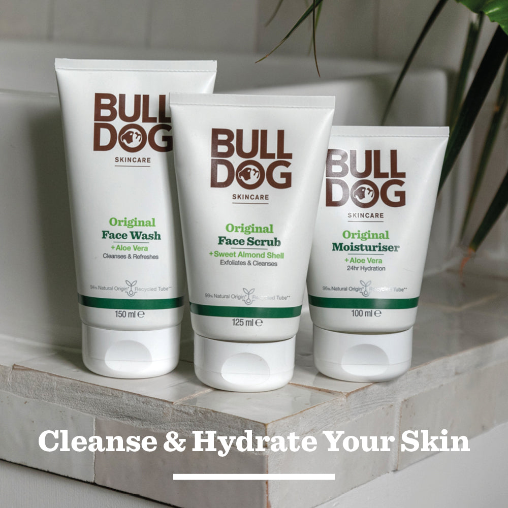 Bulldog Skincares Original Face Wash, face scrub, and moisturizer with natural ingredients are displayed on a bathroom counter beside a plant. At the bottom, text reads: Cleanse & Hydrate Your Skin.