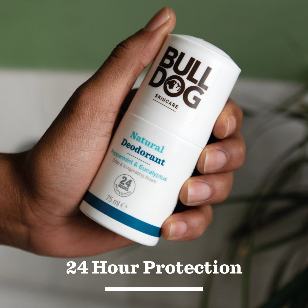 A hand holds a stick of Peppermint & Eucalyptus Natural Deodorant for men with prebiotic from chicory root, shown against a blurred background. Label highlights 24-hour odour protection.