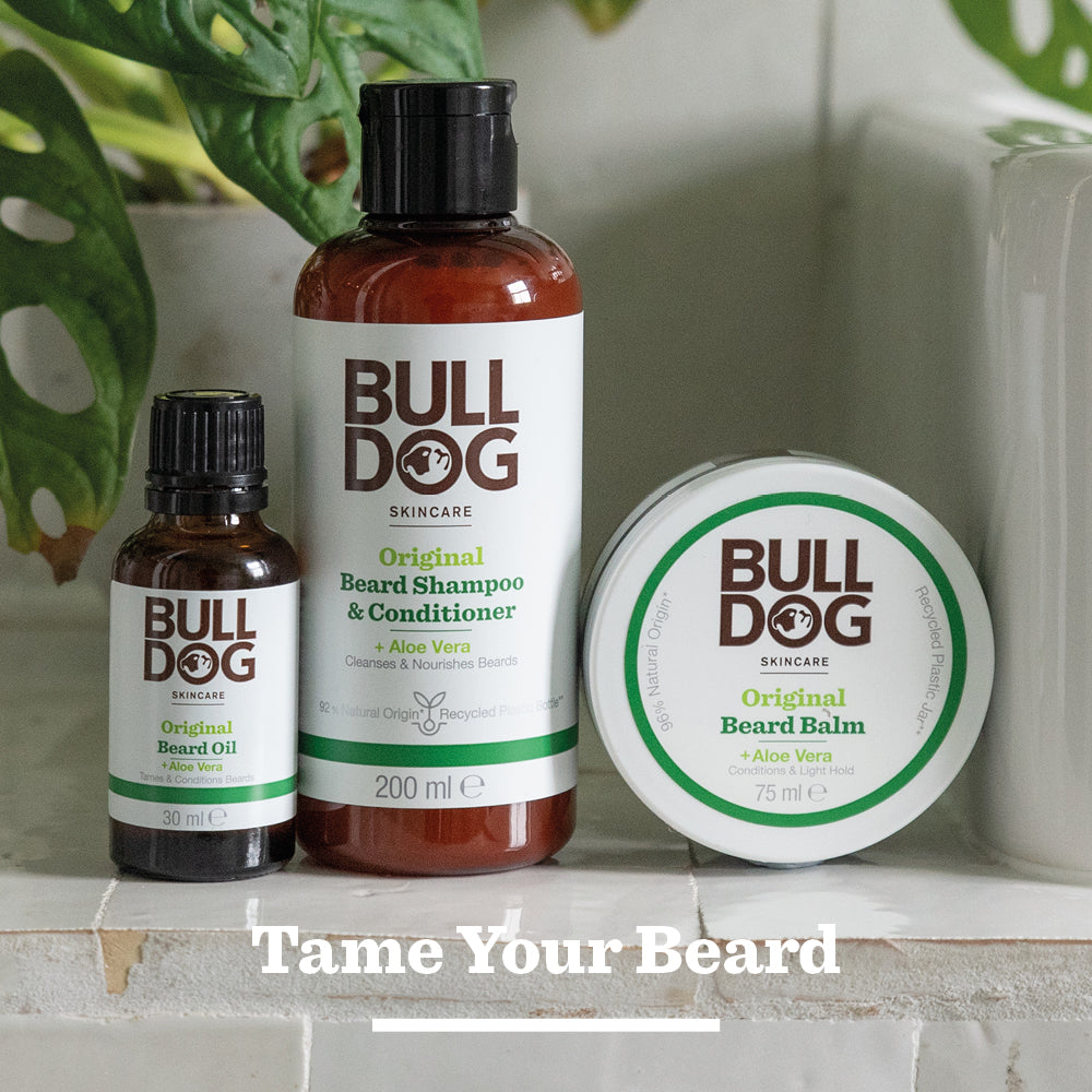 Bulldog Skincares Original Beard Wax—made with moisturising, natural origin ingredients—is displayed on a surface among green plants, with the text Tame Your Beard at the bottom.