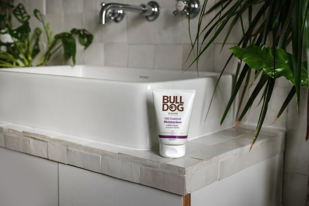 A tube of Oil Control Moisturiser rests on the rim of a white bathroom sink, surrounded by green plants and white tiled walls, highlighting this moisturiser as an ideal choice for oil-prone skin.