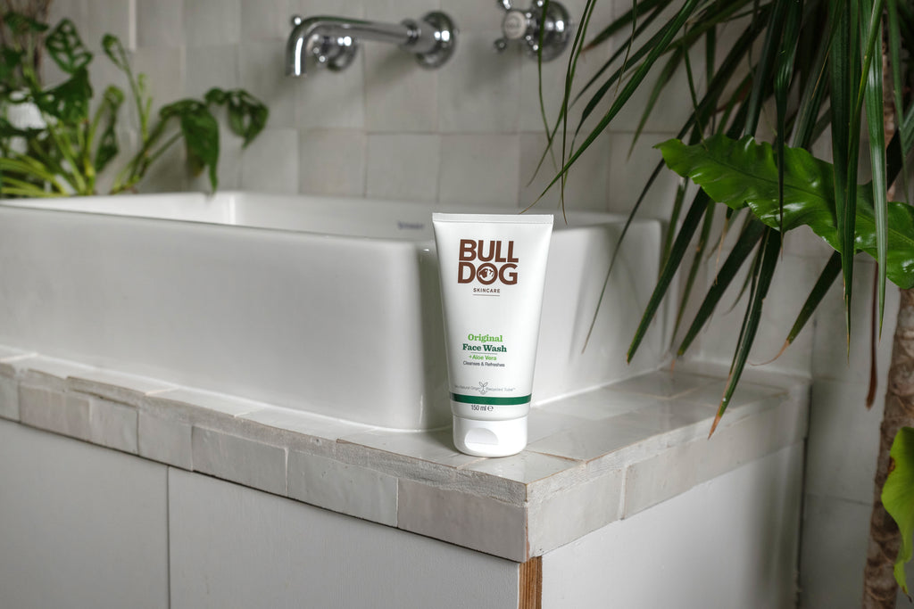 A tube of Original Face Wash sits on the edge of a white bathroom sink, surrounded by green leafy plants and white tiled surfaces.