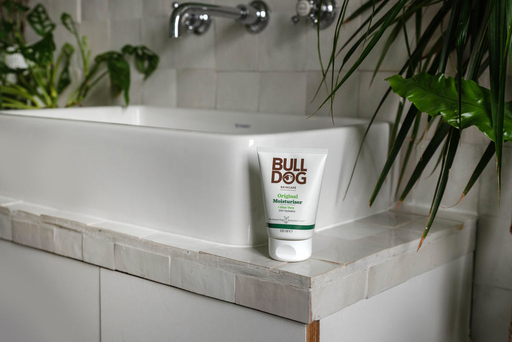 A tube of Original Moisturiser sits on the edge of a white bathroom sink, framed by green houseplants and tiled surfaces.