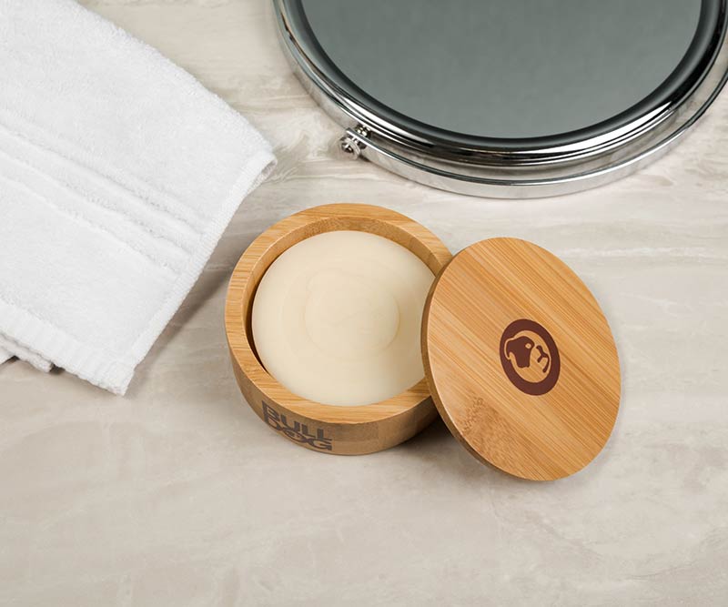 The Original Shave Soap, a refillable round bar in an open bamboo container with lid, sits on light marble beside a folded white towel and circular mirror—perfect for an eco-friendly shave.