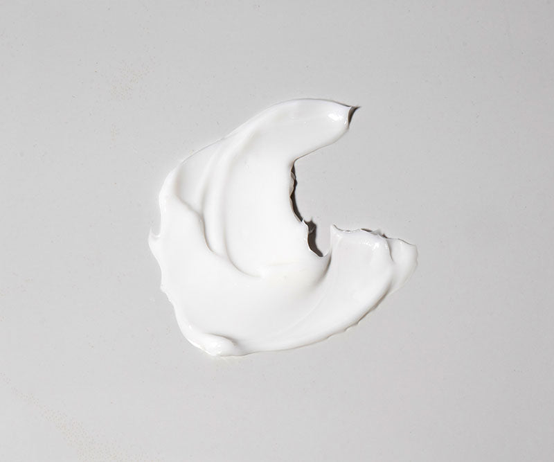 A dollop of Age Defence Moisturiser, enriched with naturally derived ingredients, is smeared in a crescent shape on a smooth, light gray surface.